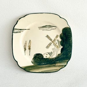 May include: A square ceramic plate with rounded corners featuring a dark green transferware print on a cream background. The design depicts a landscape with a windmill, a house, two sailboats on water, and dense foliage along the right side.