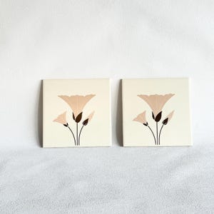 May include: Two square ceramic tiles featuring a cream-colored background with a centered graphic illustration of a stylized flower in shades of light pink, dark brown, and black. Each tile displays the same floral motif with thin stems and petals.