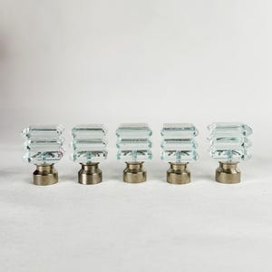 May include: Five clear, faceted glass curtain rod finials with a rectangular design. Each finial sits atop a brushed metal base. The finials are arranged in a row against a white background.