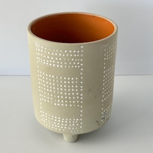 May include: A cylindrical ceramic planter with a light beige exterior featuring a white dotted grid pattern. The interior of the vessel is a vibrant orange. The planter is supported by three small, integrated feet at the base.