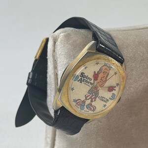 May include: Vintage gold-toned watch with a black leather band. The watch face features a cartoon illustration of a man in patriotic attire, with the text "Spiro Agnew Original" and "Dirty Time Company". The watch is Swiss made.