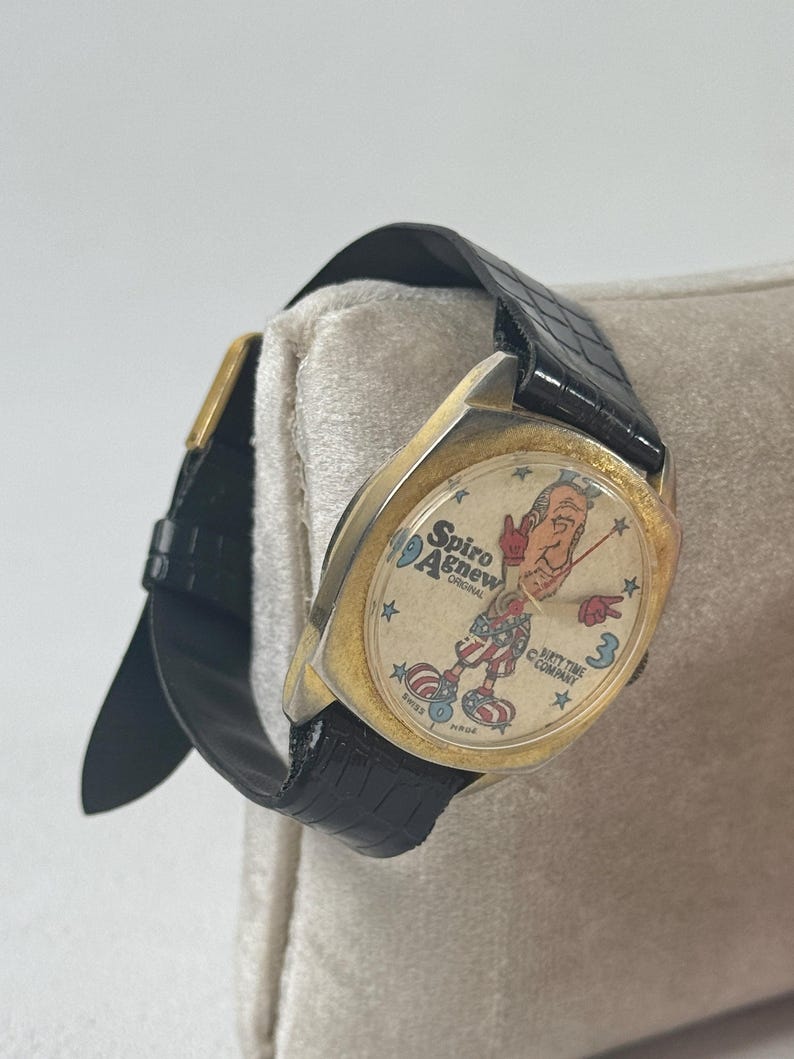 May include: Vintage gold-toned watch with a black leather band. The watch face features a cartoon illustration of a man in patriotic attire, with the text "Spiro Agnew Original" and "Dirty Time Company". The watch is Swiss made.