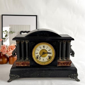 May include: An antique black mantel clock with a gold-toned clock face. The clock features Roman numerals, decorative columns, and ornate detailing. The clock's base has decorative feet and a marble-like accent.