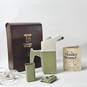 May include: A vintage Stanley Steamer travel garment steamer in olive green and white, with a brown carrying case. Includes accessories and a product information booklet. The steamer is designed to remove wrinkles from clothing.
