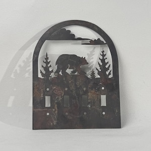 May include: Metal light switch cover featuring a bear walking across a mountain range with pine trees and clouds. The cover has three switch openings and a rustic, brown finish.