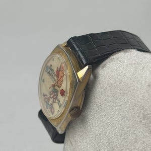 May include: Vintage gold-toned watch with a black leather band. The watch face features a colorful illustration with text. The watch has a round face and a textured black band. The watch is a classic accessory.