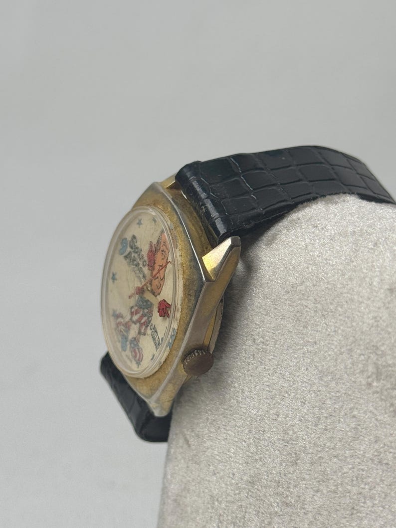 May include: Vintage gold-toned watch with a black leather band. The watch face features a colorful illustration with text. The watch has a round face and a textured black band. The watch is a classic accessory.