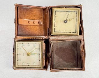 May include: An open, vintage brown leather travel case with two square clocks. The clocks have gold-toned faces, black numerals, and gold hands. The case is open, revealing the clocks and a compartment.