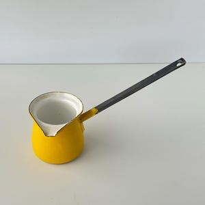 May include: A bright yellow enamel coffee pot with a white interior and a long, dark gray metal handle. The pot features a distinct pouring spout and a rounded base, with visible wear along the rim and handle attachment point.