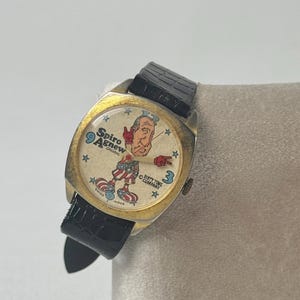 May include: Vintage gold-toned watch with a black leather band. The watch face features a cartoon illustration of a man with the text "Spiro Agnew Original" and "Sixty Time Company". The watch hands point to 9 and 3.