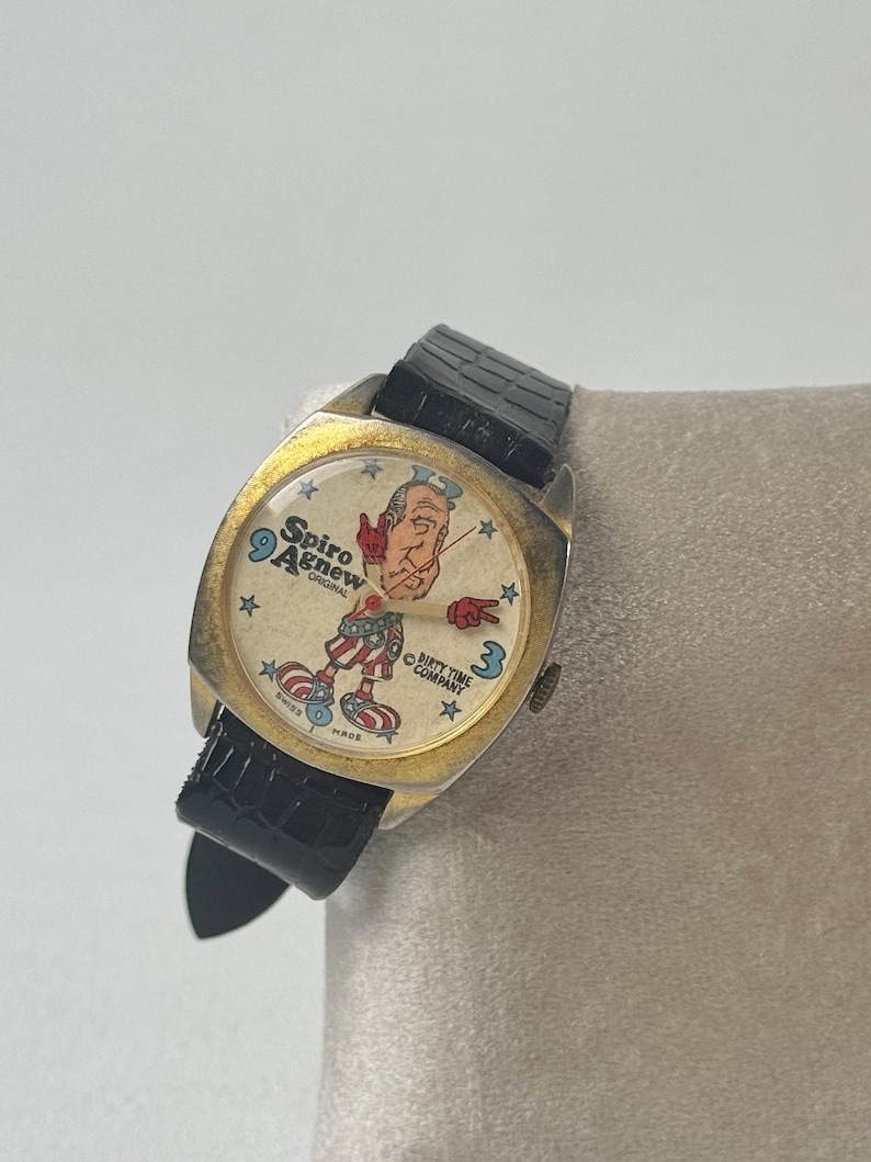 May include: Vintage gold-toned watch with a black leather band. The watch face features a cartoon illustration of a man with the text "Spiro Agnew Original" and "Sixty Time Company". The watch hands point to 9 and 3.