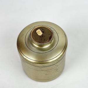 May include: A cylindrical brass container with a lid. The lid features a small, circular portrait of a person in a dark coat. The container has a slightly aged appearance, with visible wear and tear.