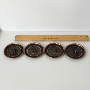 May include: Four circular metal coasters with a dark bronze finish and a raised letter D in the center. A wooden ruler is positioned behind the coasters, showing they are approximately 3 inches in diameter each, with a raised rim edge.