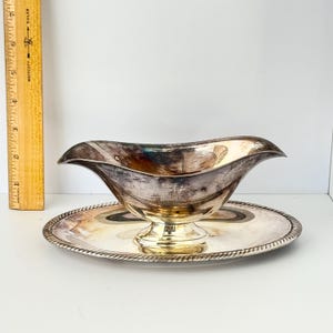 May include: A silver-toned gravy boat with a pedestal base and a matching oval underplate featuring a rope-style edge. A wooden ruler is positioned vertically next to the item, showing the gravy boat stands approximately 3.5 inches tall.