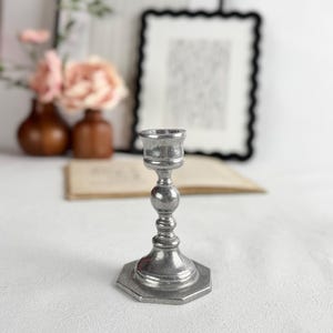 May include: A small, silver-colored candlestick holder with a hexagonal base. The candlestick has a detailed, ornate design with a round center and a cup-shaped top. The background includes blurred decor and a white surface.