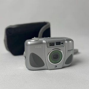 May include: A silver Kodak Advantix C750 camera with a green lens and a black carrying case. The camera has a sleek design with gray accents and the Kodak logo. The carrying case is black and has a gray strap.