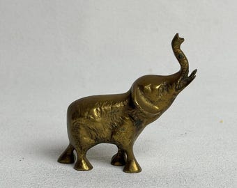 May include: A small, vintage brass elephant figurine with its trunk raised. The elephant has a textured surface and is standing on four small legs. The figurine is a warm, golden color and appears to be made of solid brass.