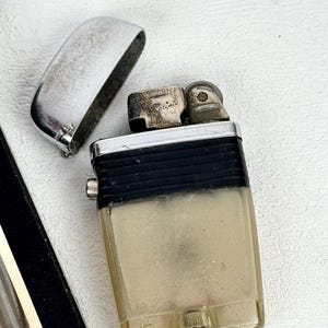 May include: Vintage Scripto lighter with a silver flip-top lid and a clear, rectangular body. The lighter has a black band and a metal spark wheel. The text "Scripto" is visible on the lighter.