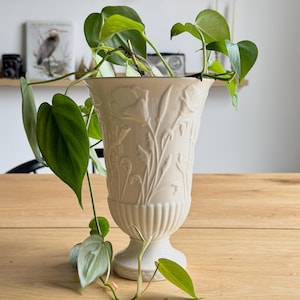 May include: A cream-colored, embossed urn-shaped vase with a floral design, holding a trailing green plant. The vase sits on a light-colored wooden table. The plant has heart-shaped leaves.