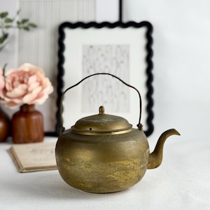 May include: A vintage brass teapot with a curved spout, a lid, and a handle. The teapot has a rounded body and a tarnished, aged appearance. The background includes framed artwork and floral arrangements, suggesting a decorative or antique theme.