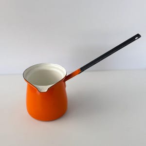May include: A bright orange enamel coffee pot with a white interior and a long, slender black handle. The pot features a small pouring spout on the rim, a rounded base, and a smooth, glossy finish throughout the exterior surface of the vessel.