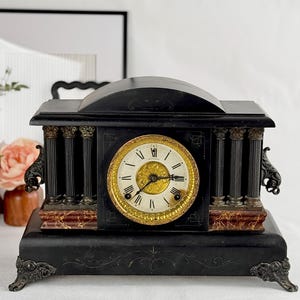 May include: An antique black mantel clock with a round clock face featuring gold accents and Roman numerals. The clock has decorative columns and ornate details, with a marble-like base. The clock is a vintage home decor item.
