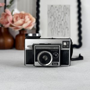 May include: A vintage Instamatic 414 camera with a black and silver finish. The camera has the Kodak logo and the text "Instamatic 414 Camera". The camera is on a white surface.