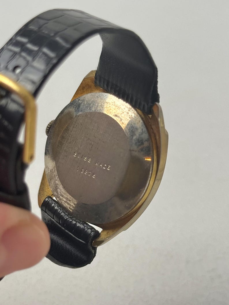 May include: Vintage gold-toned wristwatch with a black leather band. The watch face is not visible, but the back of the watch is visible, with the words "SWISS MADE" and the number "13826" engraved on the metal casing.