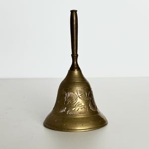 May include: A vintage brass bell with a dark brown handle. The bell has an etched floral design around the base. The bell is sitting on a white surface.