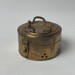 May include: A small, round, antique brass box with a hinged lid and a handle. The box has a latch closure and decorative cross-shaped cutouts. The brass has a warm, aged patina.