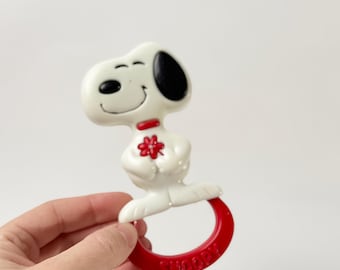 May include: A white Snoopy baby rattle with a red collar and flower detail. The rattle has a red ring base with the word "SNOOPY" printed on it. The Snoopy figure has a black ear and is smiling. The rattle is held by a hand.