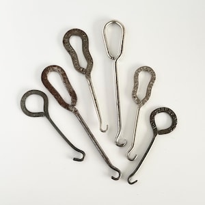 May include: A collection of five vintage metal button hooks with varying handle shapes and finishes. Some handles feature embossed text including Economy Shoe, Walk-Over Shoes, and Bond Street, while others have textured patterns or smooth surfaces.