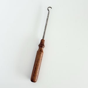 May include: A metal button hook tool featuring a long, slender, silver-toned steel shaft with a curved hook at the tip and a cylindrical, dark brown wooden handle with a smooth, tapered grip section for manual operation and handling.