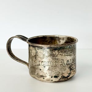 May include: A tarnished silver-toned metal mug with a curved handle. The exterior surface features a weathered, dark patina and an engraved inscription that reads Margaret Ann in a cursive script. The mug has a cylindrical body and a smooth rim.
