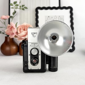 May include: Vintage Argus Seventy-five camera with a flash attachment. The camera is black and silver with the name "Argus" and "Seventy-five" printed on the front. The flash has a large, round, silver reflector. The background includes flowers and framed art.