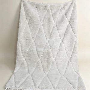May include: A rectangular off-white area rug featuring a raised diamond pattern across its surface. The rug is crafted with a thick, textured pile and finished with a row of short, knotted fringe tassels along the bottom edge of the textile.