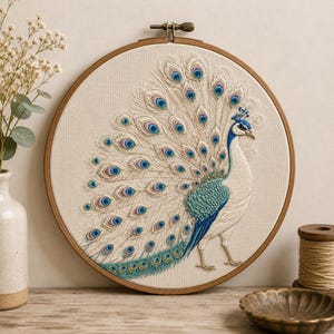 May include: A circular wooden embroidery hoop containing a detailed needlepoint design of a peacock with a fanned tail. The peacock features white, teal, and blue threadwork with intricate eye-shaped patterns in shades of purple and blue.