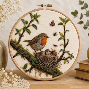 May include: A circular embroidery hoop featuring a detailed needlework scene of a robin perched on a mossy branch above a nest with two chicks. The design includes green leaves, a monarch butterfly, a bee, and a small insect on cream fabric.