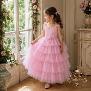 May include: A floor-length pink dress featuring a bodice with floral appliqués, thin shoulder straps, and a multi-layered tulle skirt. The dress has a sweetheart neckline and a full, voluminous silhouette. A logo for Cherie Tulle Co. is visible.