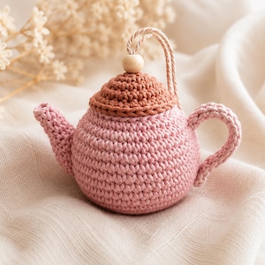 Peut inclure: Une petite théière décorative crochetée de forme ronde, réalisée en fil rose clair avec un motif texturé. Elle est dotée d'un couvercle couleur terre cuite, d'une anse et d'un bec verseur assortis, ainsi qu'une perle en bois clair sur le cordon.