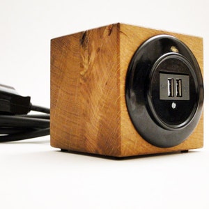 Könnte beinhalten: Ein Holzwürfel mit einer schwarzen Steckdose und zwei USB-Anschlüssen. Der Würfel ist mit einem schwarzen Stromkabel mit Stecker verbunden.