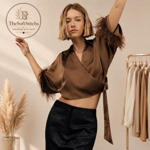 May include: A brown wrap blouse featuring a V-neckline, short sleeves with feathered trim, and a side tie closure. The garment is paired with a black skirt with a subtle tonal pattern. A circular logo in the top left corner reads TheSoftStitches.