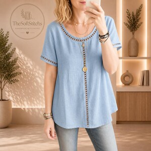 Op de afbeelding: Een lichtblauw shirt met korte mouwen en een ronde halslijn met een verticale rij cirkelvormige uitsnijdingen die in het midden naar beneden loopt. Het kledingstuk heeft bijpassende cirkelvormige uitsnijdingen langs de mouwranden.