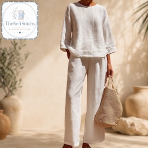 May include: A white long-sleeved top and matching wide-leg trousers set made of a textured fabric. The outfit is paired with a woven tan straw tote bag. The top features a round neckline and loose sleeves, while the trousers have a relaxed fit.