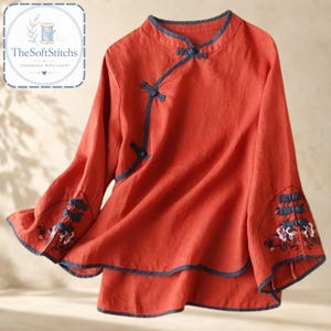 Puede incluir: Una blusa de color naranja óxido de manga larga con cuello mao, ribetes gris oscuro y cierres asimétricos de alamares en la parte delantera. Las mangas acampanadas tienen alamares decorativos gris oscuro y bordados florales en los puños.