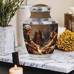 May include: A cylindrical metal cremation urn featuring a detailed wrap-around illustration of several individuals gathered around a glowing fire. The urn has a polished silver-toned lid and a matching silver-toned base with intricate engraved patterns.