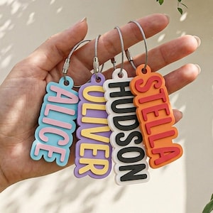 May include: Four rectangular keychains with rounded edges, each featuring raised lettering. The keychains display the names ALICE in pink on blue, OLIVER in yellow on purple, HUDSON in black on white, and STELLA in red on orange, all on metal rings.