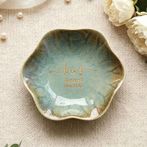 May include: A scalloped, flower-shaped ceramic trinket dish with a glossy, mottled teal and brown glaze. The center features gold-colored text that reads k & f, Engaged, and Nov. 15. 26, written in a combination of script and sans-serif fonts.