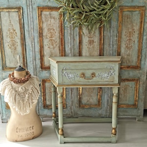 May include: A mint green side table with a single drawer, decorated with floral decals and gold accents. The table has a brass handle and stands on four legs with gold detailing. A vintage dress form and a eucalyptus wreath are in the background.
