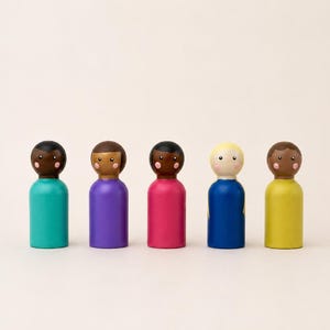 Könnte beinhalten: Fünf Holzfiguren in einer Reihe, jede mit einem individuellen Hautton und einem einfarbigen Körper. Die Figuren sind in Türkis, Lila, Magenta, Blau und Gelb bemalt und haben einfache Gesichtszüge mit Augen und rosafarbenen Wangen.